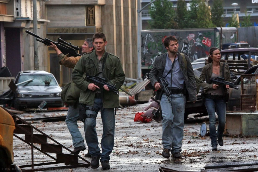 Four people with serious expressions, wearing rugged clothing and carrying guns, walk through a debris-strewn, post-apocalyptic urban street with damaged buildings, wrecked cars, and graffiti in the background.