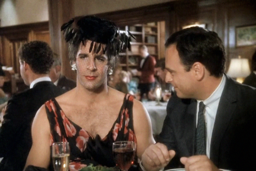 A man in a floral dress, feathered hat, and earrings sits at a dining table beside another man in a suit. They are in a restaurant with other diners and bookshelves in the background.