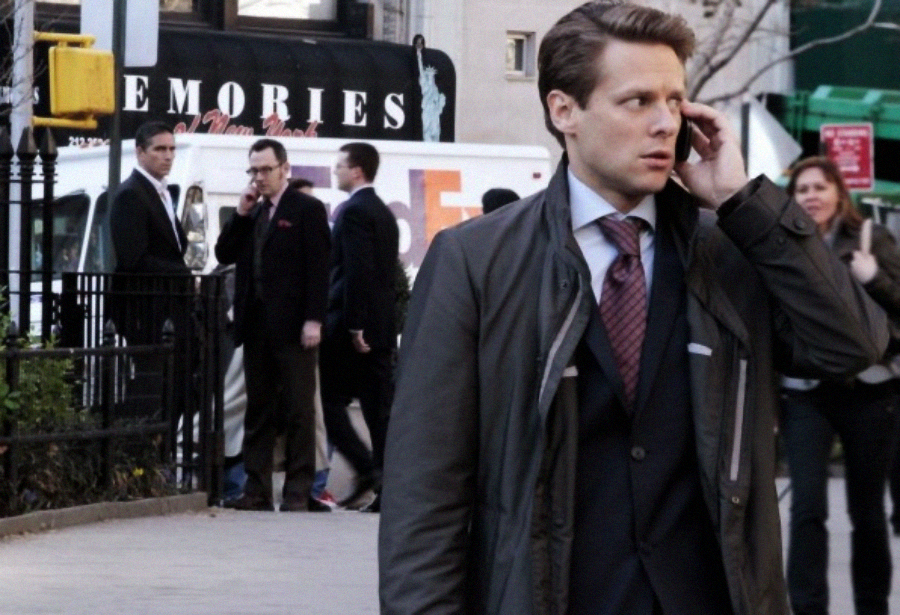 A man in a suit and coat talks on his phone while walking on a city sidewalk; several people and a white van are in the background near a cafe with a “MEMORIES” sign.