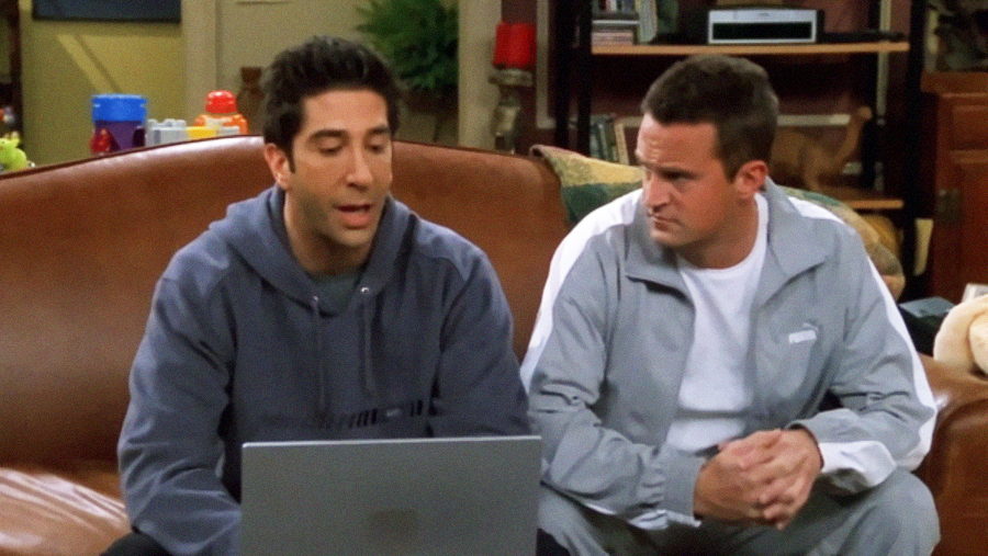 Two men sit on a brown couch, looking at a laptop. One wears a dark hoodie and is talking, while the other in a light gray tracksuit listens intently. The background shows household items and shelves.