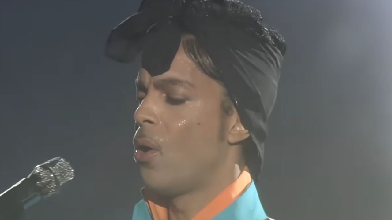 A person sings into a microphone on stage, wearing a black headwrap and a blue shirt with an orange collar, lit by stage lighting.