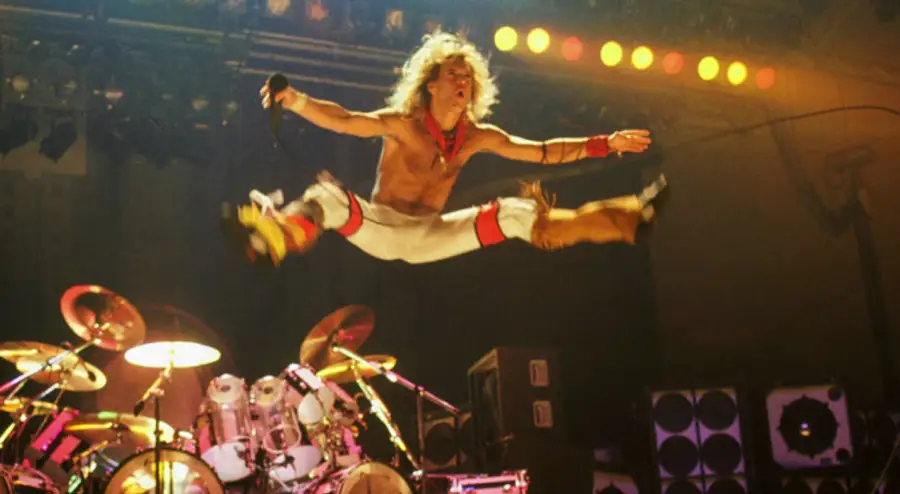 A rock singer in white pants and red accessories performs a mid-air split jump on stage, holding a microphone, with drum set and amplifiers visible in the background under stage lights.
