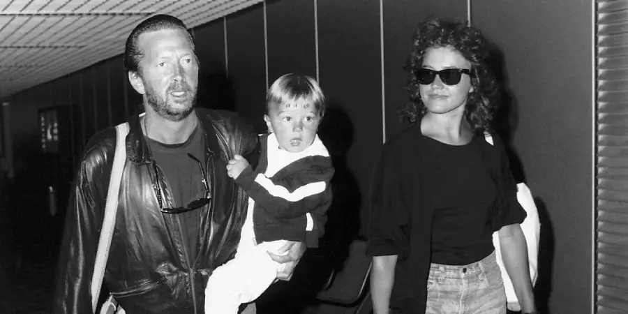 A man wearing a leather jacket carries a young boy while walking beside a woman in sunglasses and casual clothes. They are indoors, possibly at an airport. The image is in black and white.