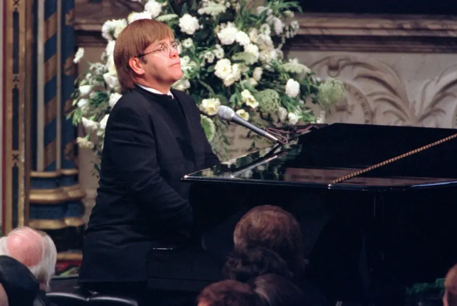 A person in a black suit and glasses plays a grand piano in front of an audience, with a large bouquet of white flowers in the background.