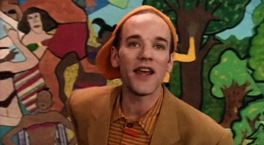 A person wearing an orange beret and striped shirt smiles in front of a colorful mural featuring abstract figures and trees.