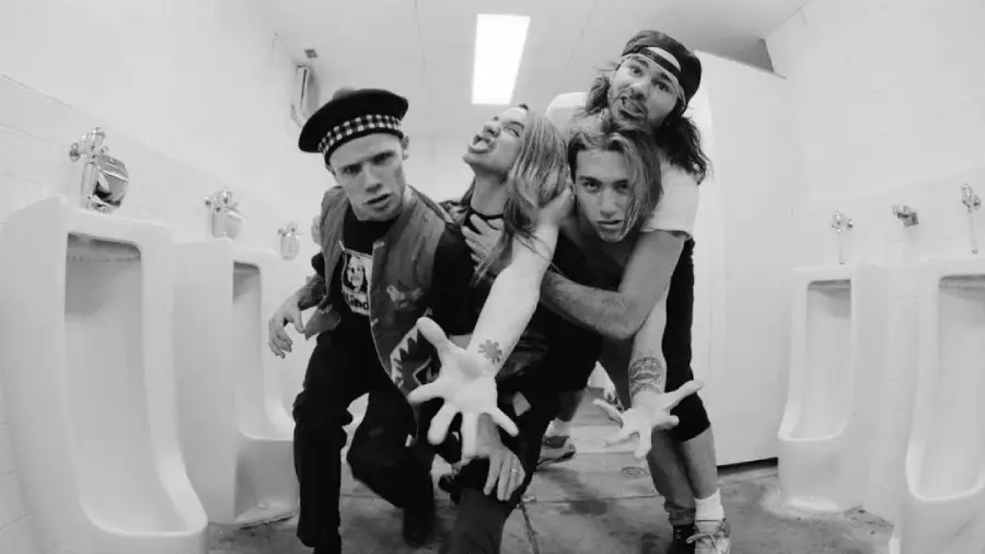 Four people pose playfully and energetically in a men's restroom with urinals visible in the background; one wears a cap, another sticks out their tongue, and all seem expressive and dynamic.