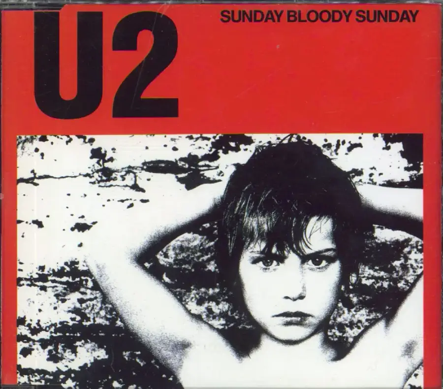 Album cover for U2’s "Sunday Bloody Sunday" featuring a black-and-white photo of a young boy with arms behind his head, set against a wall. The top is red with bold "U2" text and the song title.