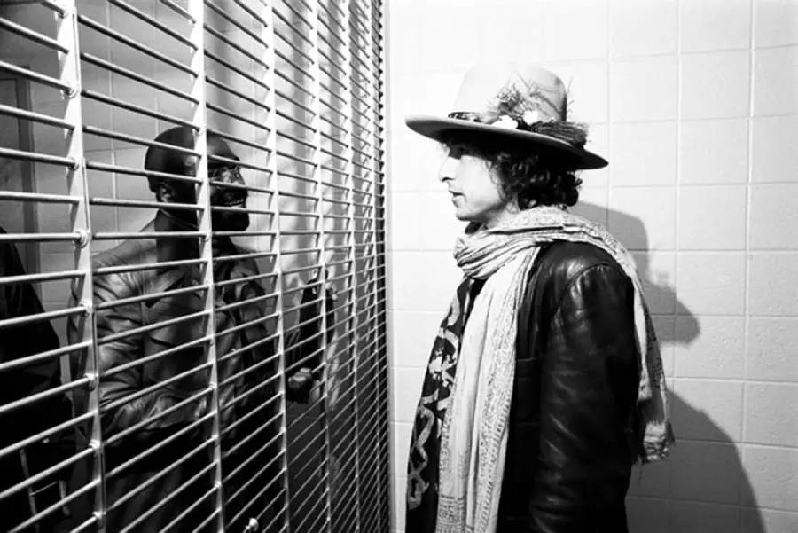 A person wearing a hat, scarf, and leather jacket stands facing another person behind metal bars in a white-tiled room. The two seem to be having a serious conversation through the barrier.