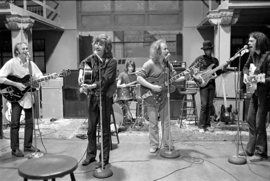 A black and white photo of a six-member band performing indoors, with five musicians playing guitars and singing into microphones, and one person playing drums in the background.