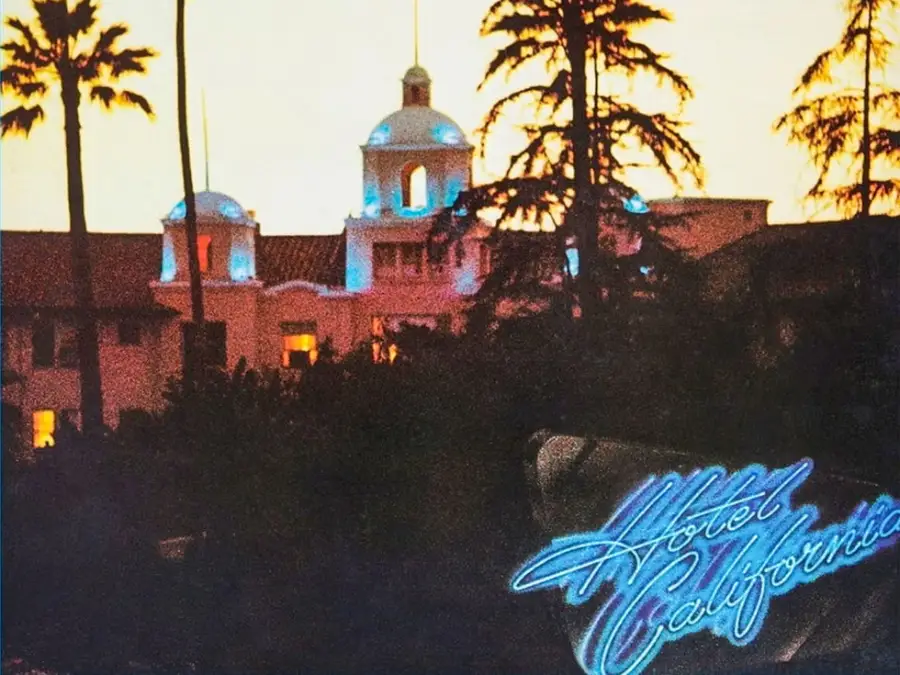A grand hotel with domed towers is silhouetted at sunset, surrounded by palm trees. The neon blue sign in the foreground reads “Hotel California.”