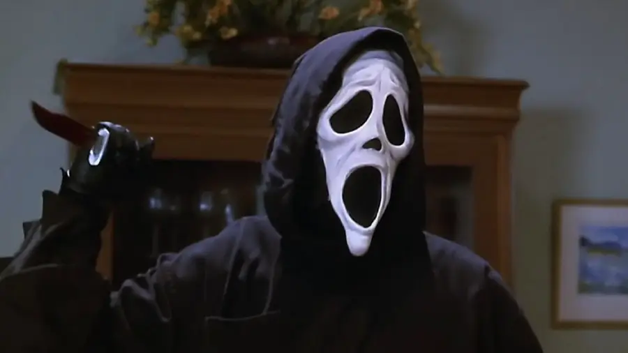A person wearing a black hooded cloak and a white, elongated ghost mask holds a large knife raised in their gloved hand, standing indoors with shelves and a painting in the background.
