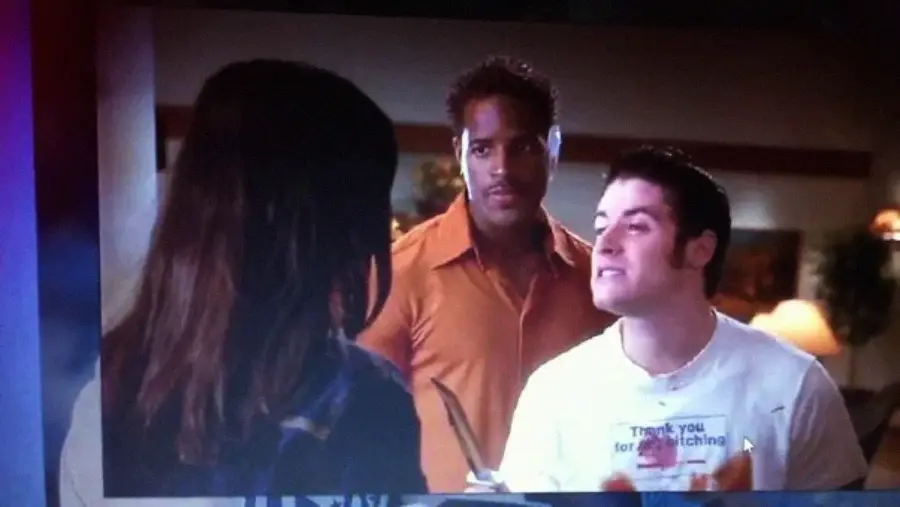 A scene from a TV show or movie featuring three people indoors; a woman with long dark hair faces two men, one wearing an orange shirt and the other in a white shirt, who appears to be angrily holding an object.