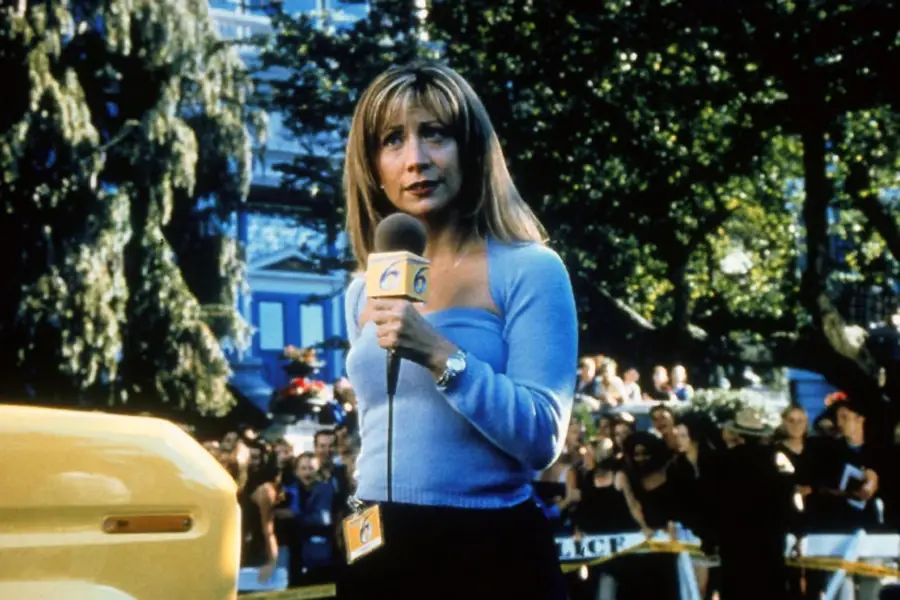 A woman with blonde hair holds a microphone and speaks on camera outdoors, with a crowd and police tape in the background on a sunny day.