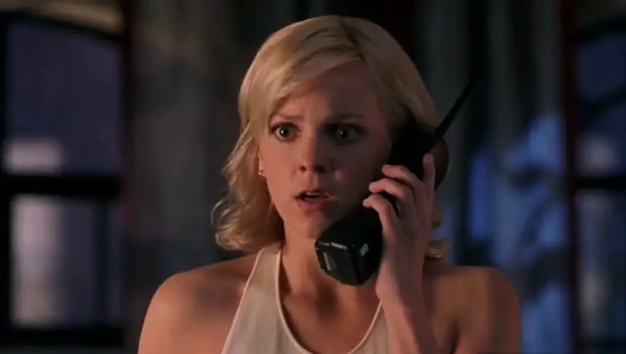 A blonde woman with a shocked expression holds a black cordless phone to her ear, wearing a white halter top, with blurred windows in the background.