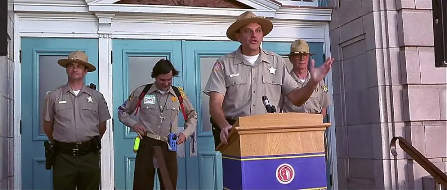 Four uniformed officers stand in front of blue doors; one is speaking at a podium, while another adjusts his belt. The setting appears to be a formal or official event outside a building.