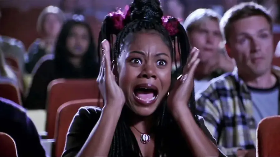 A woman sitting in a theater looks shocked and frightened, holding her hands to the sides of her face with her mouth open, while people around her react with surprise.