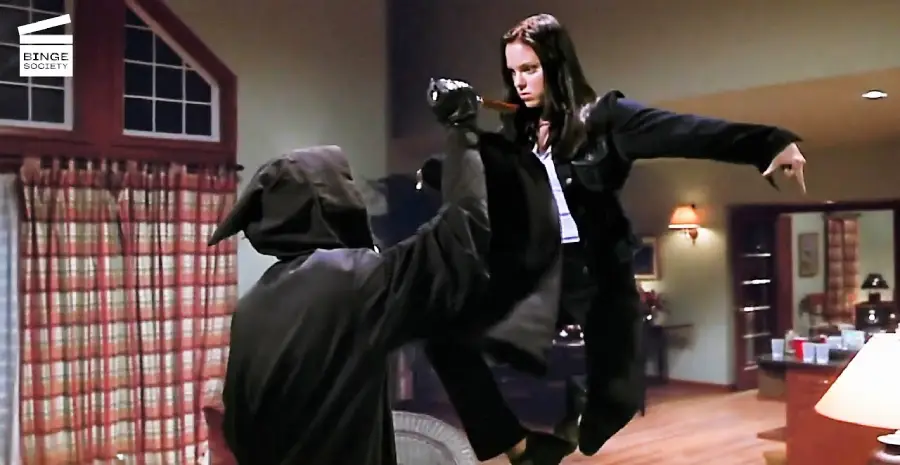 A woman in black kicks a hooded figure in a living room, appearing mid-air during the action scene. The room has large windows with curtains, warm lighting, and furniture in the background.