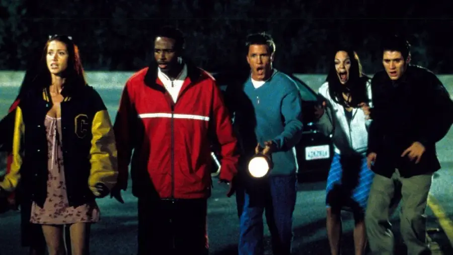 Five young adults stand close together at night, looking frightened. One holds a flashlight, and a car is visible behind them. Their expressions suggest fear or shock, and they appear to be in a tense situation outdoors.