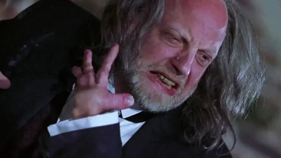 A disheveled man with long gray hair and a beard grimaces in pain or anger, baring his teeth and raising a gnarled hand with twisted fingers. He wears a suit and is illuminated by dramatic lighting.