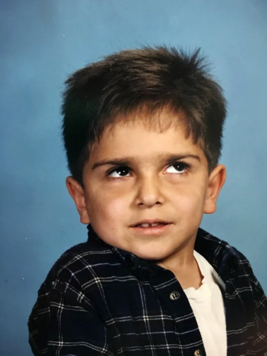 A young boy with short brown hair wearing a plaid shirt over a white T-shirt looks slightly upward and to the side with a playful, mischievous expression against a blue background.