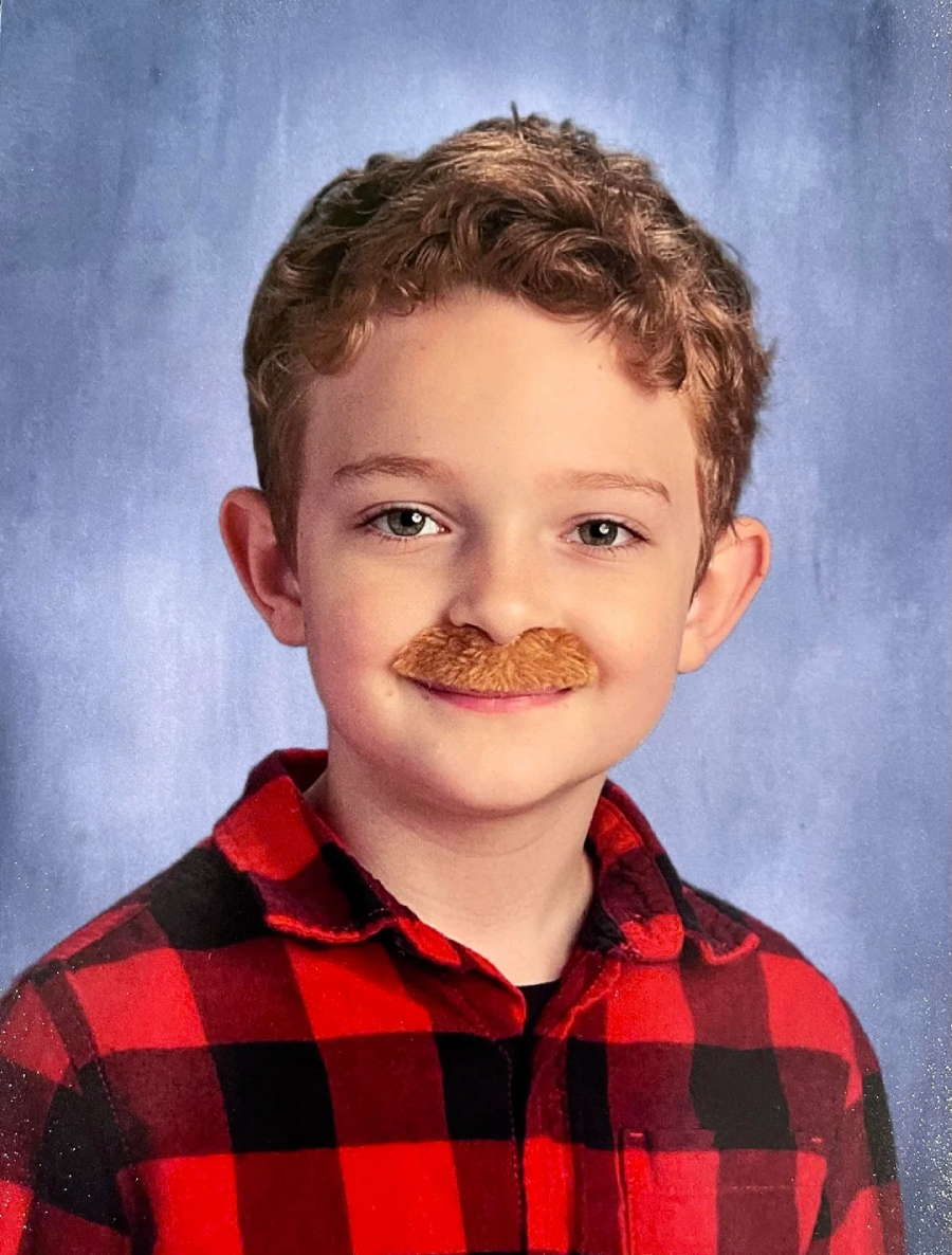 A young boy with curly hair and blue eyes wears a red and black plaid shirt, smiling at the camera. He has a fake brown mustache, and the background is a soft, blue studio-style backdrop.