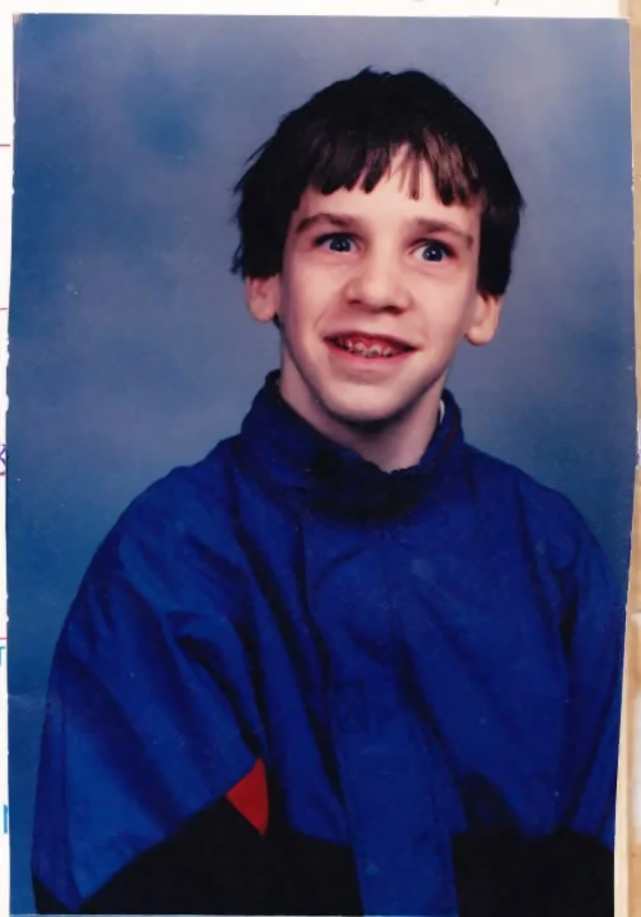 A young person with short brown hair, wearing a blue jacket and smiling with visible braces, poses against a blue studio background.