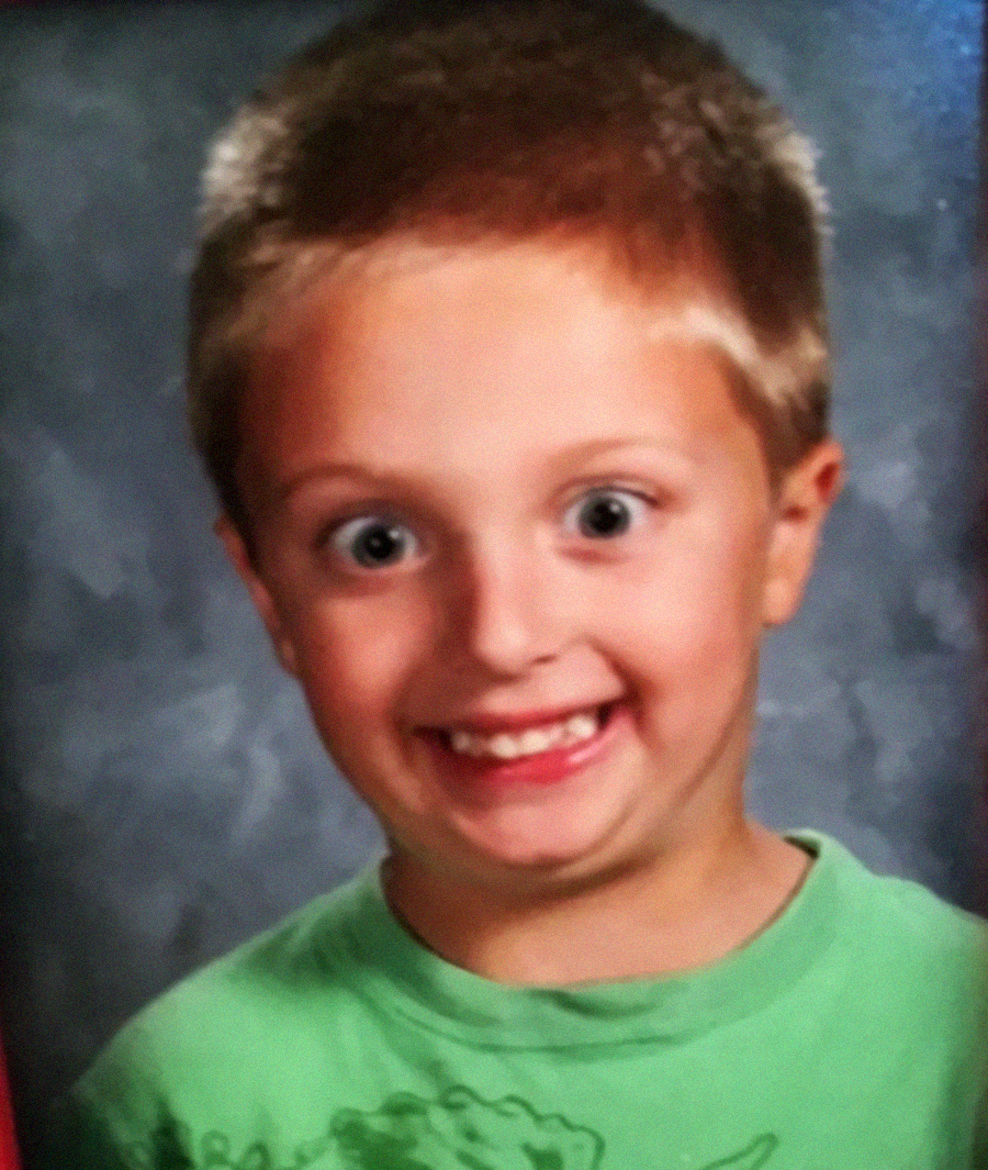 A young boy with short light brown hair and blue eyes smiles widely at the camera, wearing a bright green shirt. The background is a neutral gray, suggesting a school photo.