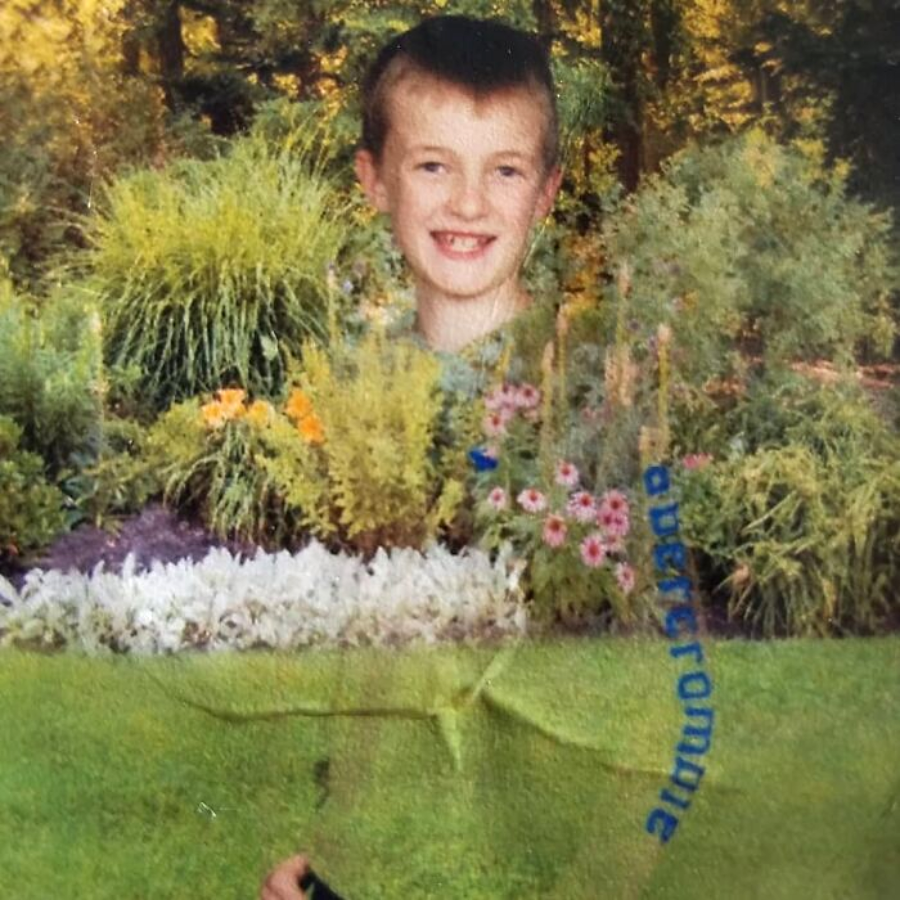 A boy is smiling in front of a lush garden, but his body is partially invisible, blending into the background, likely due to a photographic or printing error.