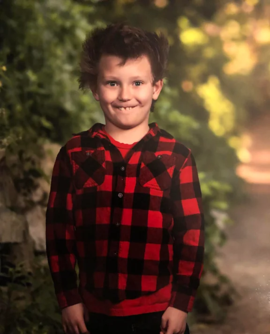 A young child with tousled hair smiles playfully at the camera, wearing a red and black plaid shirt. The background is outdoors with greenery and a softly lit path.