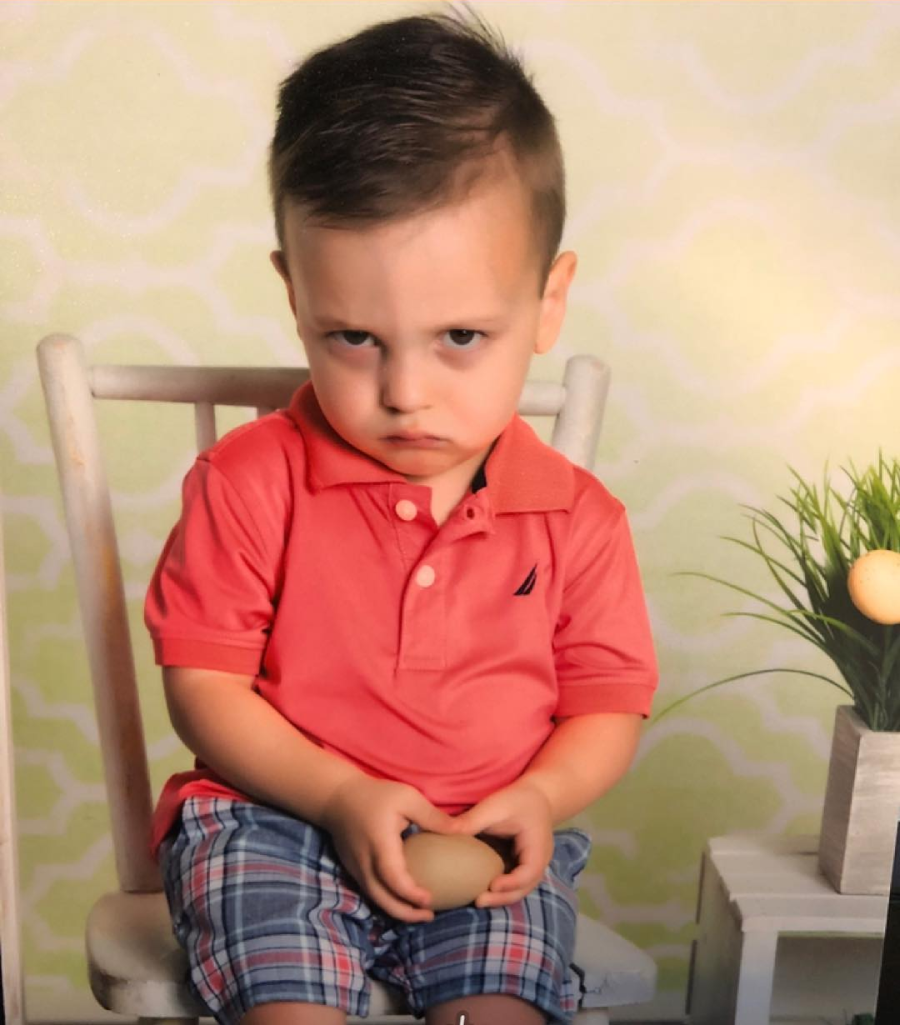 A young boy sits on a white chair, wearing a coral polo shirt and plaid shorts, looking grumpy with a frown. He holds an egg and sits beside a small table with a potted plant.
