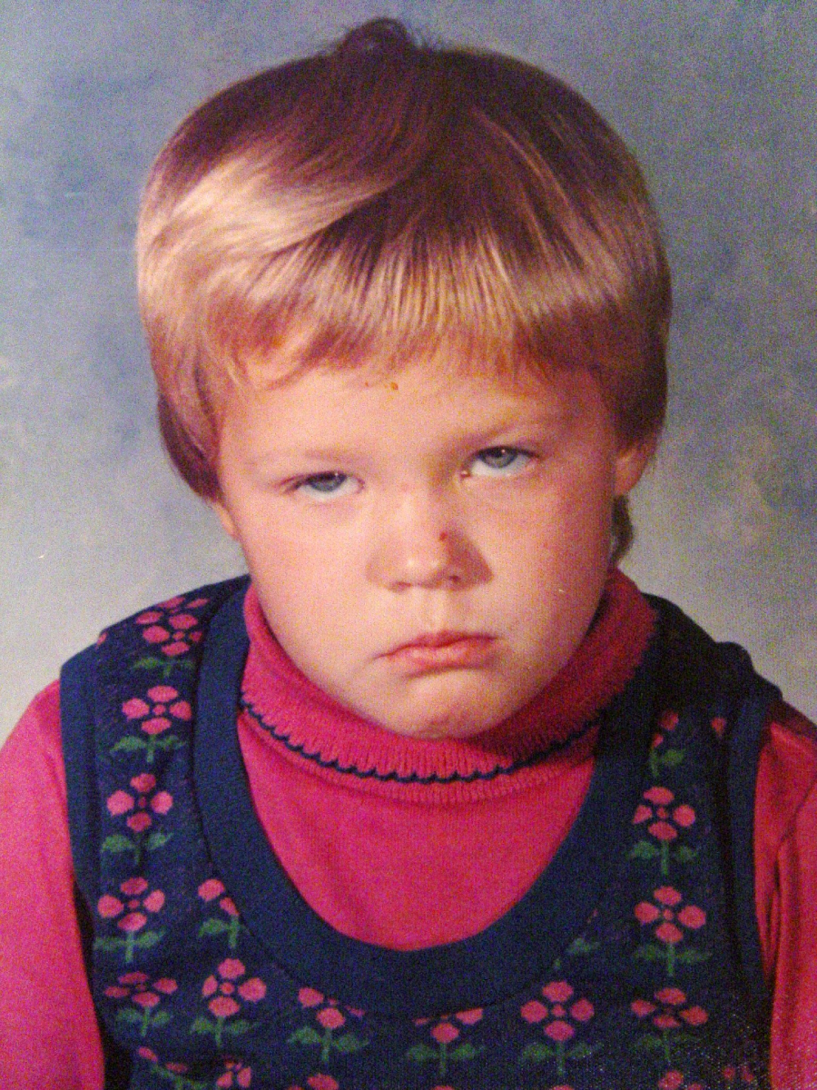 A young child with short, light brown hair wears a pink turtleneck and a navy blue dress with a pink floral pattern, looking directly at the camera with a serious, slightly unhappy expression.