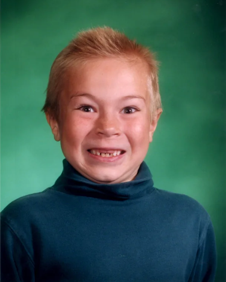 A young child with short blond hair smiles widely, showing missing teeth, and wears a teal turtleneck against a bright green studio background.