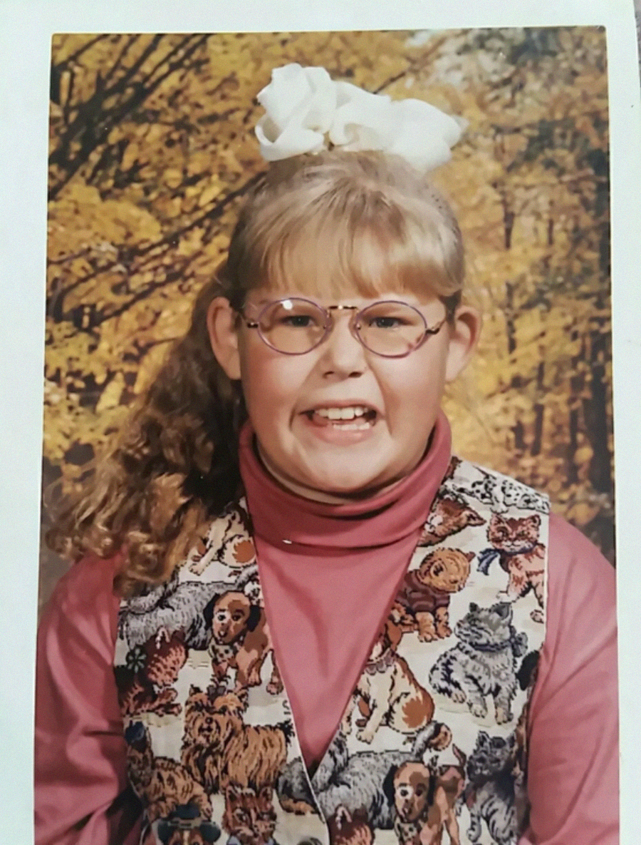 A young girl with blonde hair, a large white bow, and round glasses smiles widely. She wears a pink turtleneck and a vest decorated with dog illustrations, with a fall foliage background.
