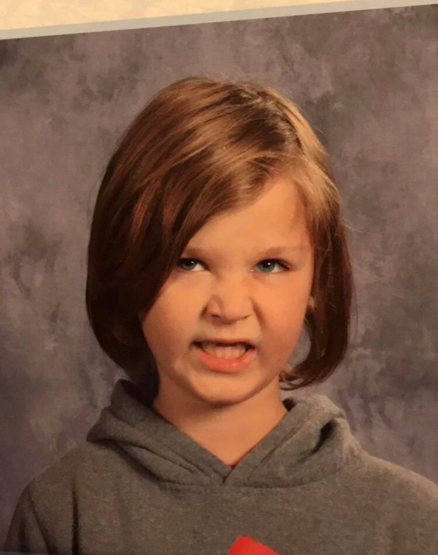 A young child with shoulder-length brown hair and a gray hoodie makes a playful, scrunched-up face, baring their teeth slightly, in front of a mottled gray backdrop.