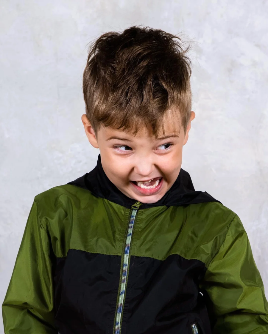 Young boy with light brown hair wearing a green and black jacket, looking to the side and making a playful, mischievous facial expression with his mouth open and eyes wide. Light, neutral background.