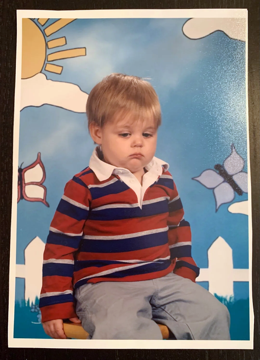 A young child with blonde hair, wearing a red, blue, and white striped shirt and gray pants, sits looking sad in front of a colorful backdrop with a sun, clouds, butterflies, and a white picket fence.