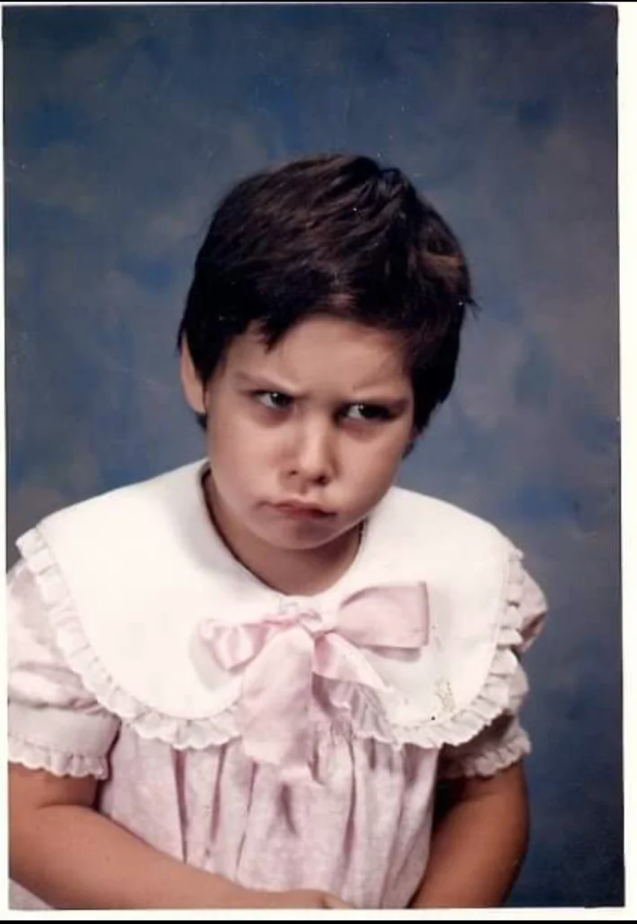 A young child with short dark hair, wearing a pink dress with a large white collar and pink bow, looks up with a pouty, grumpy expression against a blue mottled background.
