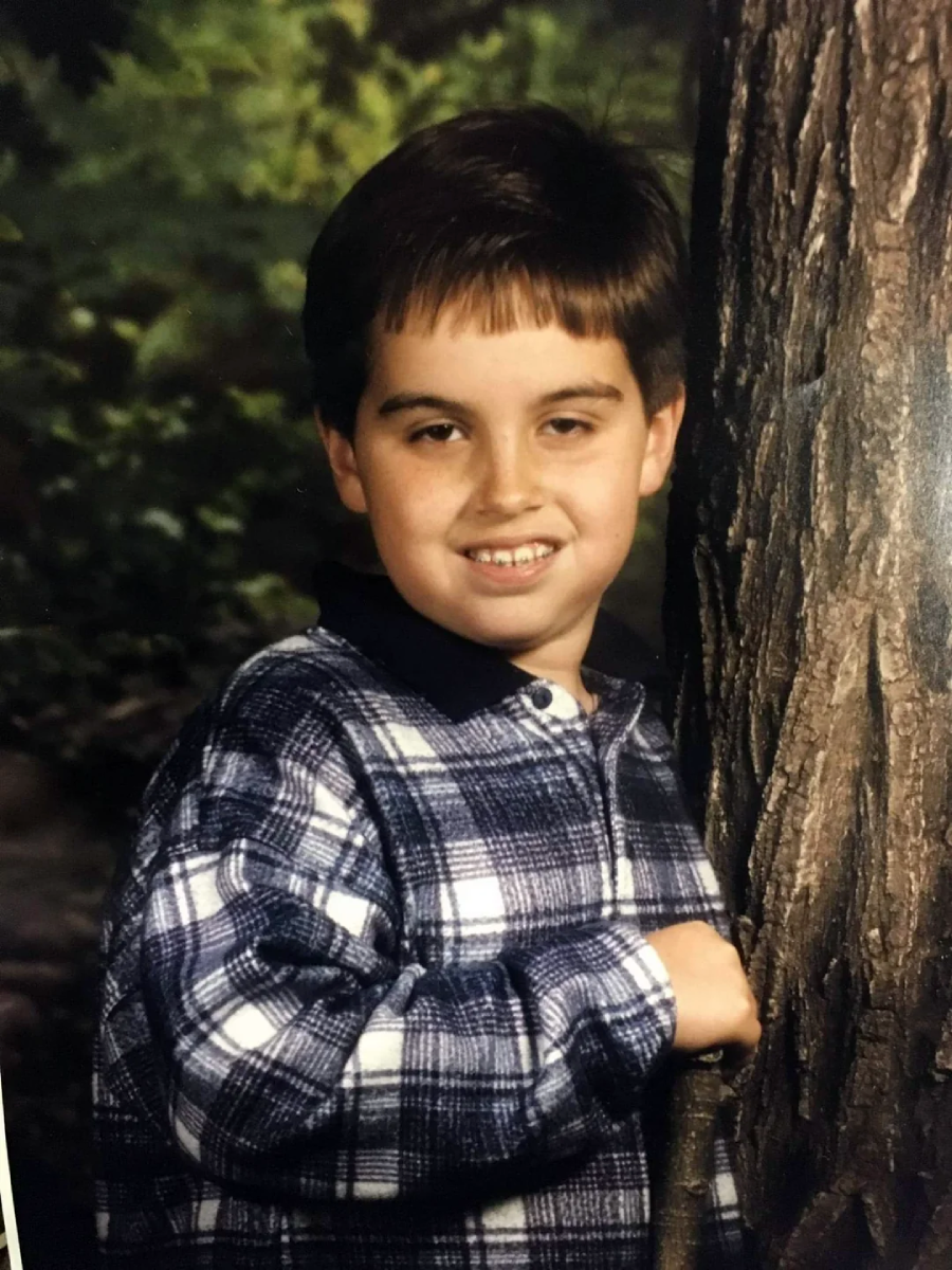 A young boy with short brown hair and a plaid shirt smiles while standing next to a tree, with a blurred green background suggesting an outdoor setting.