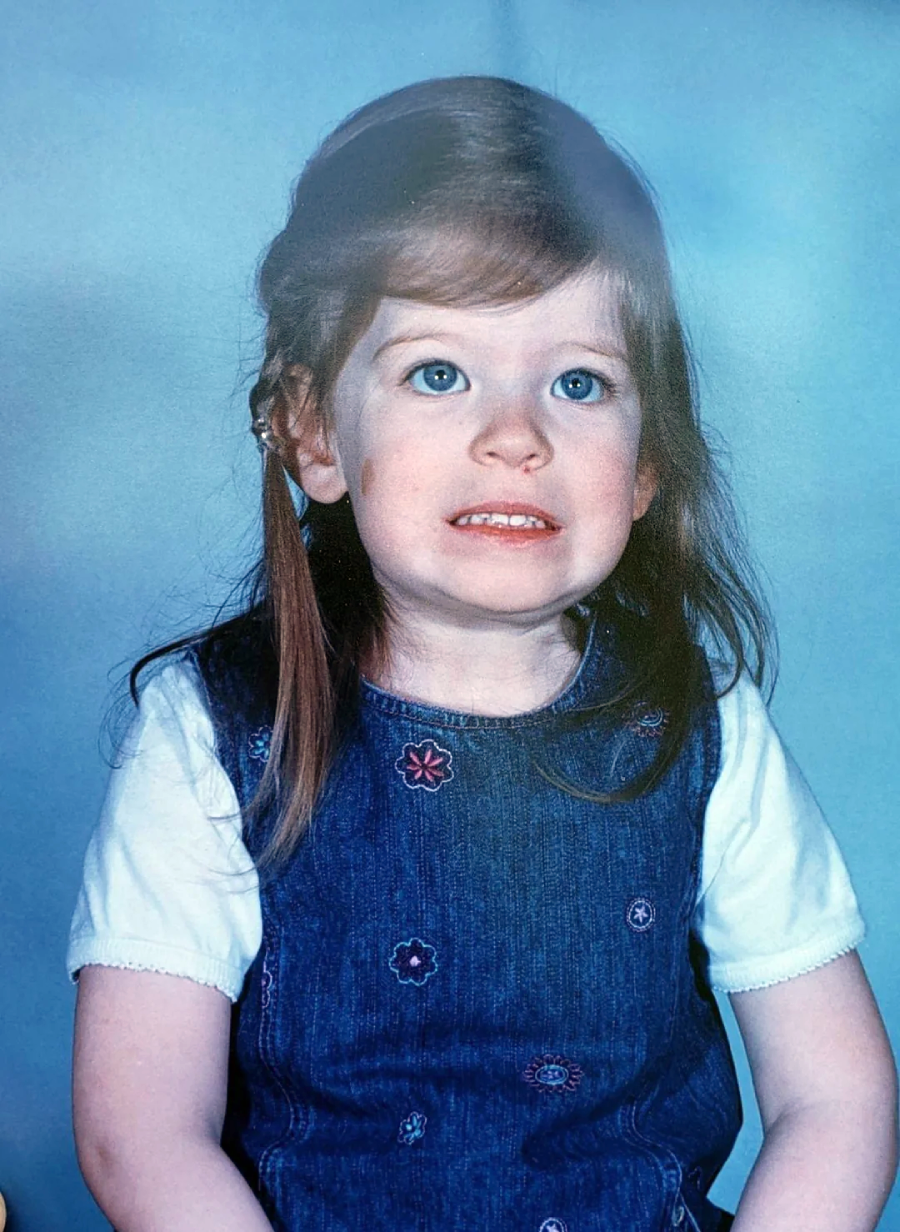 A young girl with blue eyes and long brown hair, wearing a white shirt under a blue denim dress, sits in front of a blue background and smiles awkwardly with her teeth clenched.