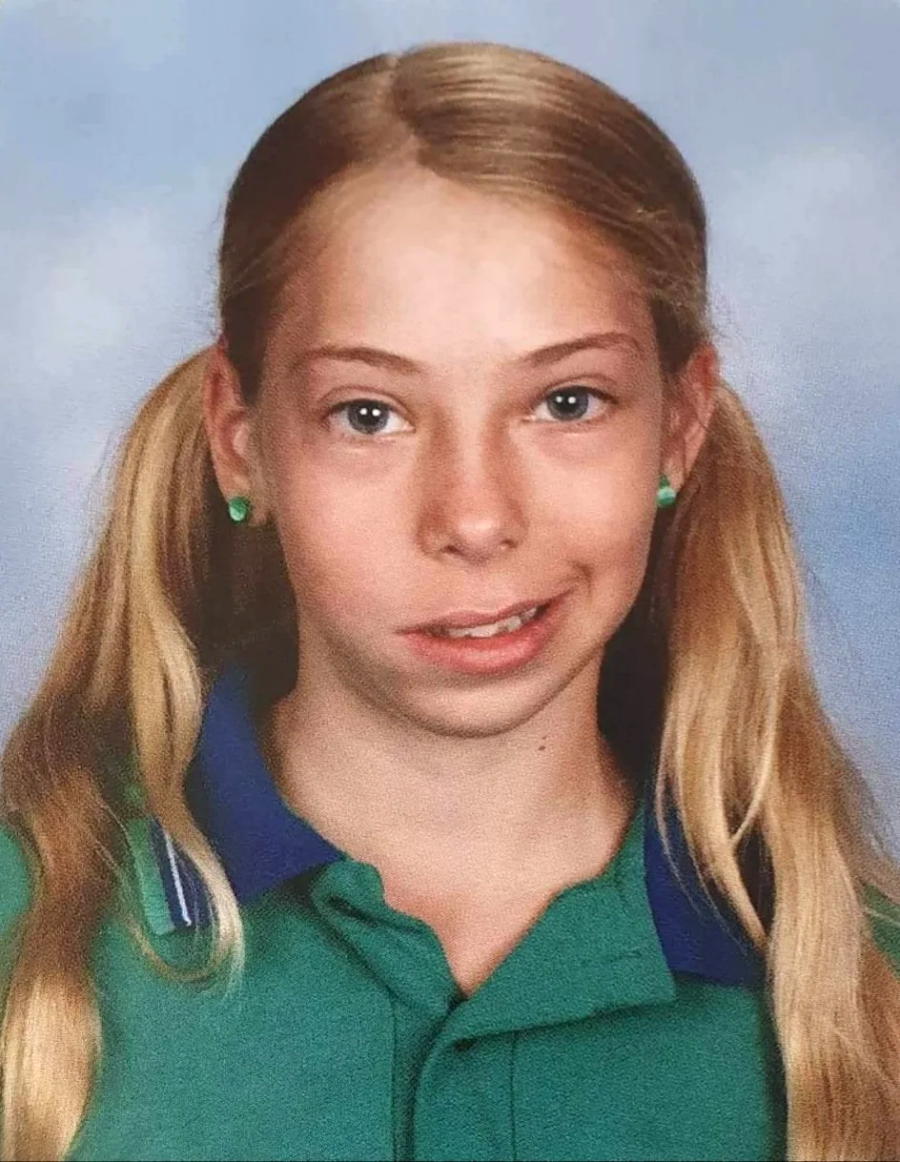 A young girl with long blonde hair in pigtails, wearing green earrings and a green collared shirt with blue trim, poses for a school photo against a light blue background, giving an awkward, uneven smile.