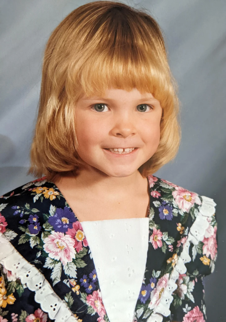 A young child with blond hair and bangs smiles at the camera, wearing a floral dress with a white collar, set against a soft, blue-gray portrait background.