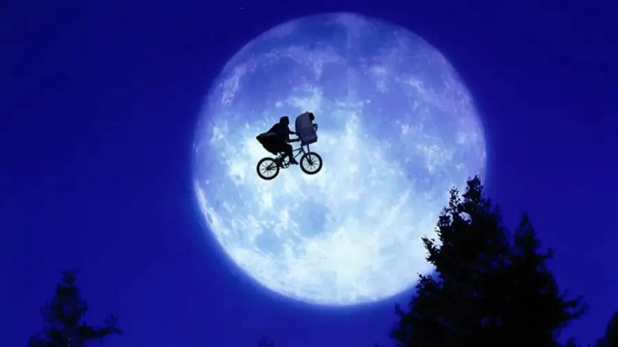 A silhouette of a child riding a bicycle with a figure in the front basket, flying across a large, bright full moon in a deep blue night sky, with dark tree tops visible below.