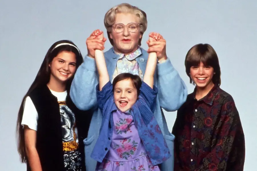 A person dressed as an elderly woman stands behind three smiling children, holding up the hands of the youngest girl in the center. All four are posing together against a plain background.