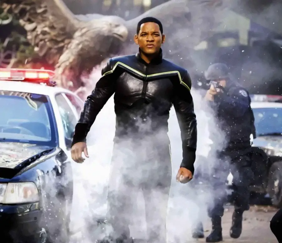 A man in a black superhero suit stands confidently between two damaged police cars, surrounded by smoke, with police officers and a large eagle statue in the background.