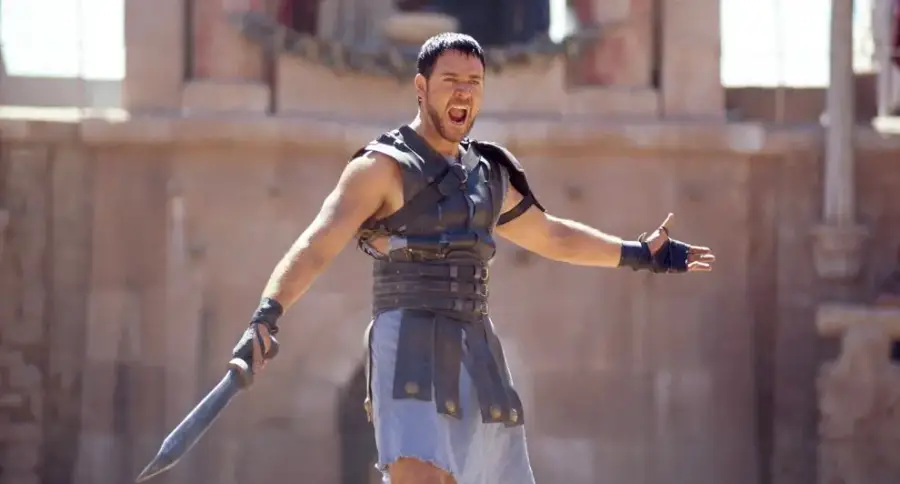 A man in Roman gladiator armor stands in an arena with his arms outstretched, holding a sword and shouting, with a determined expression on his face.