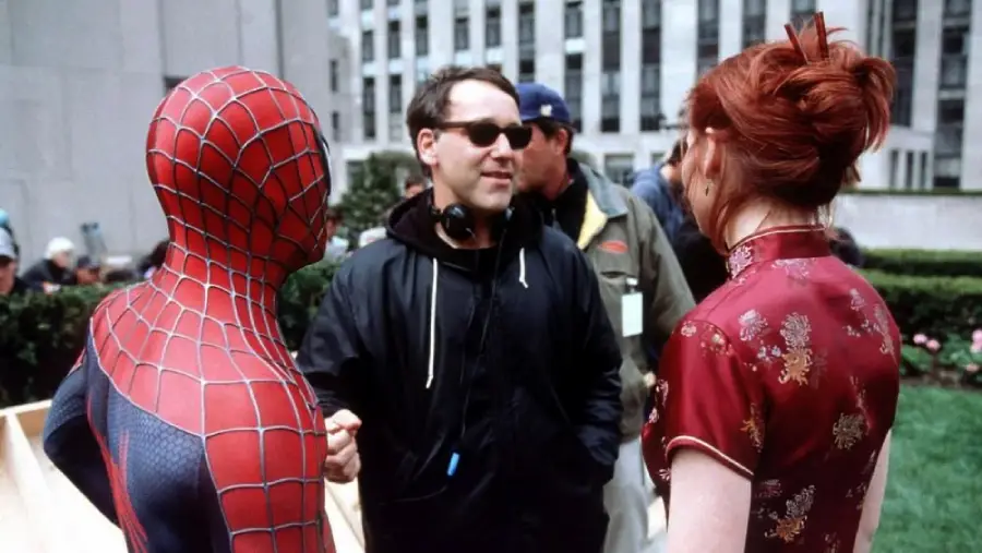 A person in a Spider-Man costume, a man in sunglasses and a headset, and a woman with red hair in a red dress talk outdoors in a city garden setting.