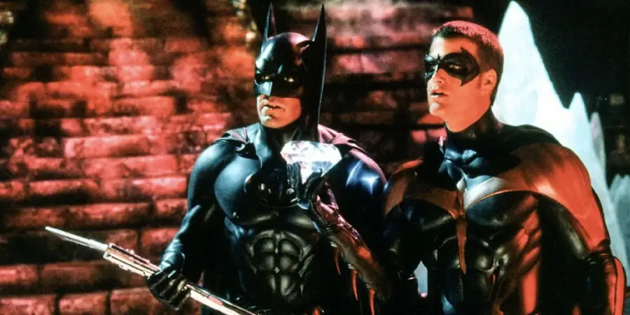 Batman and Robin, dressed in their black and red superhero costumes, stand side by side in a dimly lit, cave-like setting. Robin holds a large, glowing diamond while Batman holds a staff.