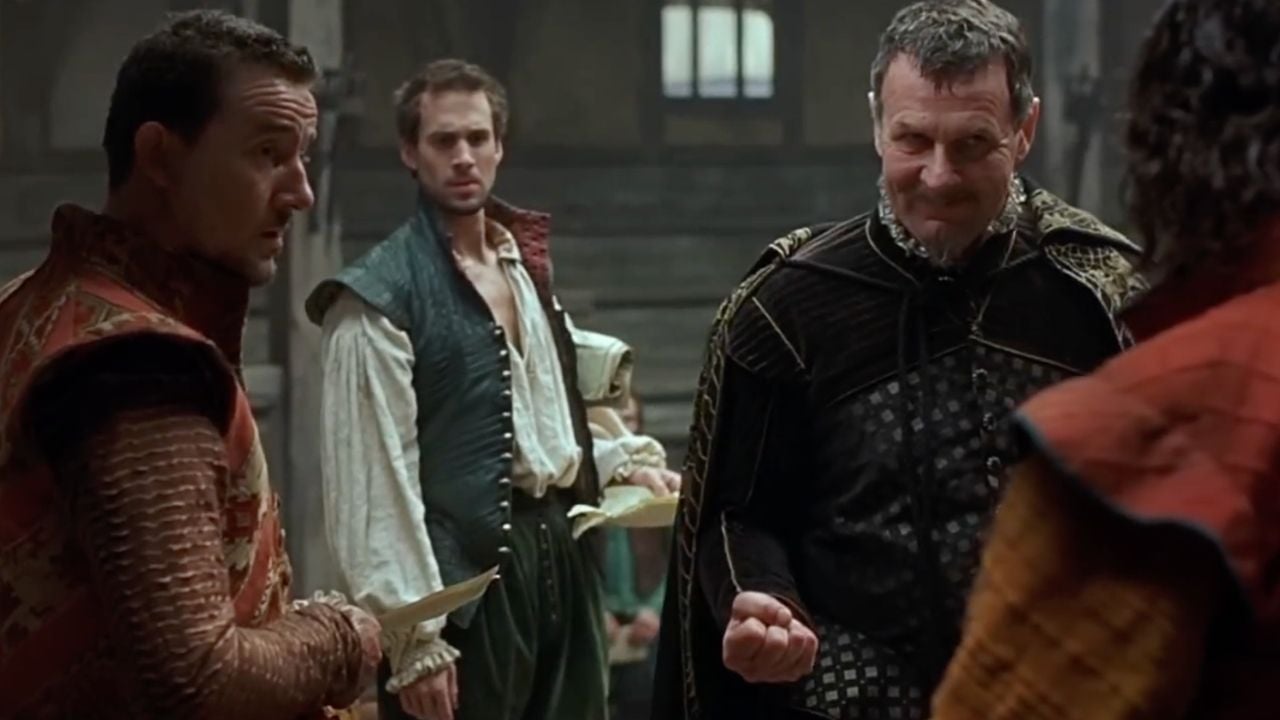 Three men in Renaissance costumes have a tense conversation indoors; one man stands in the background holding papers, while another in dark clothing clenches his fist and smirks.