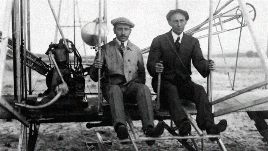 Two men in early 20th-century clothing sit side by side on the frame of a vintage biplane, surrounded by exposed machinery and open landscape in the background.