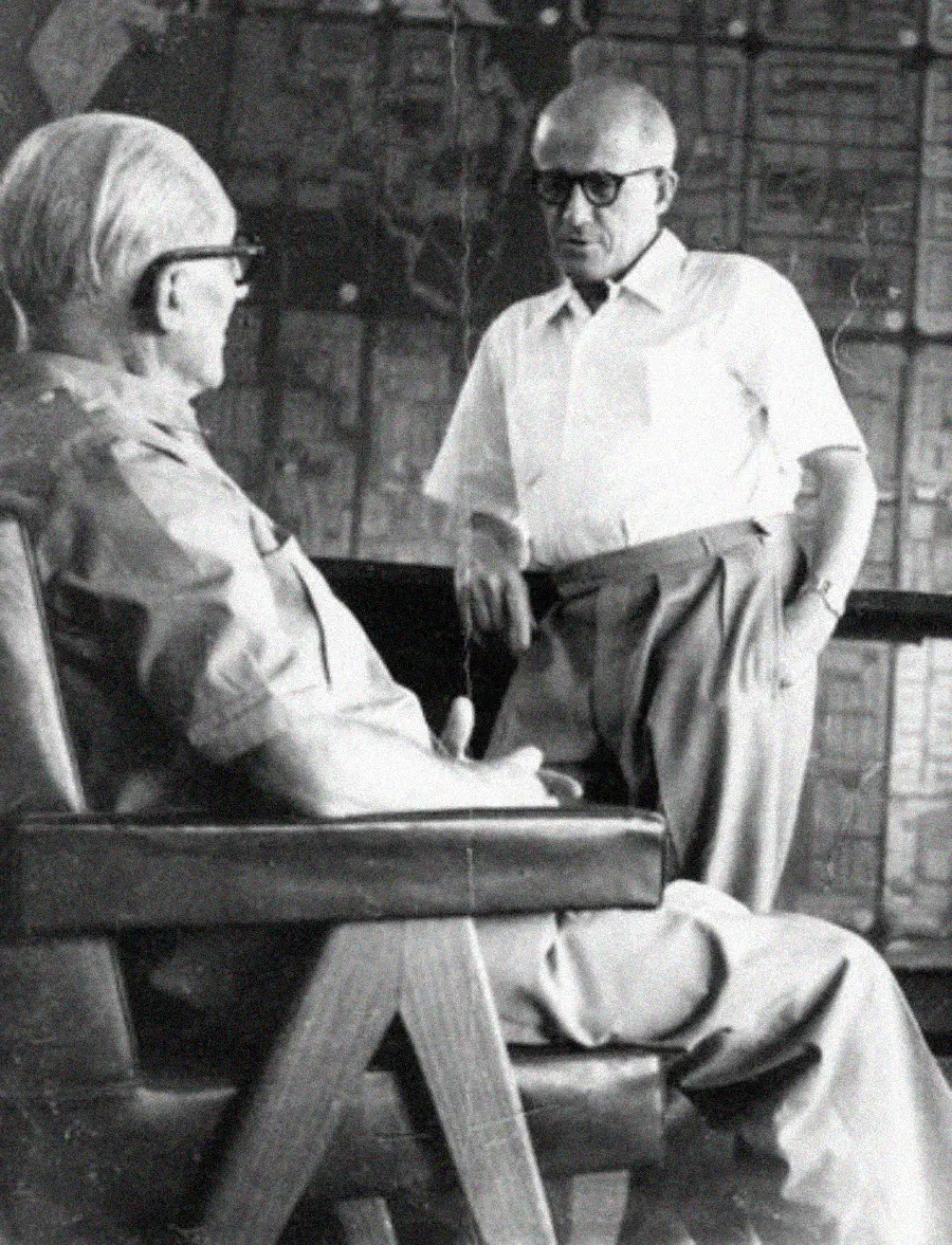 Two older men with glasses have a conversation; one is seated in an armchair facing partially away, while the other stands, leaning on the chair. A large map or architectural plan is visible in the background.